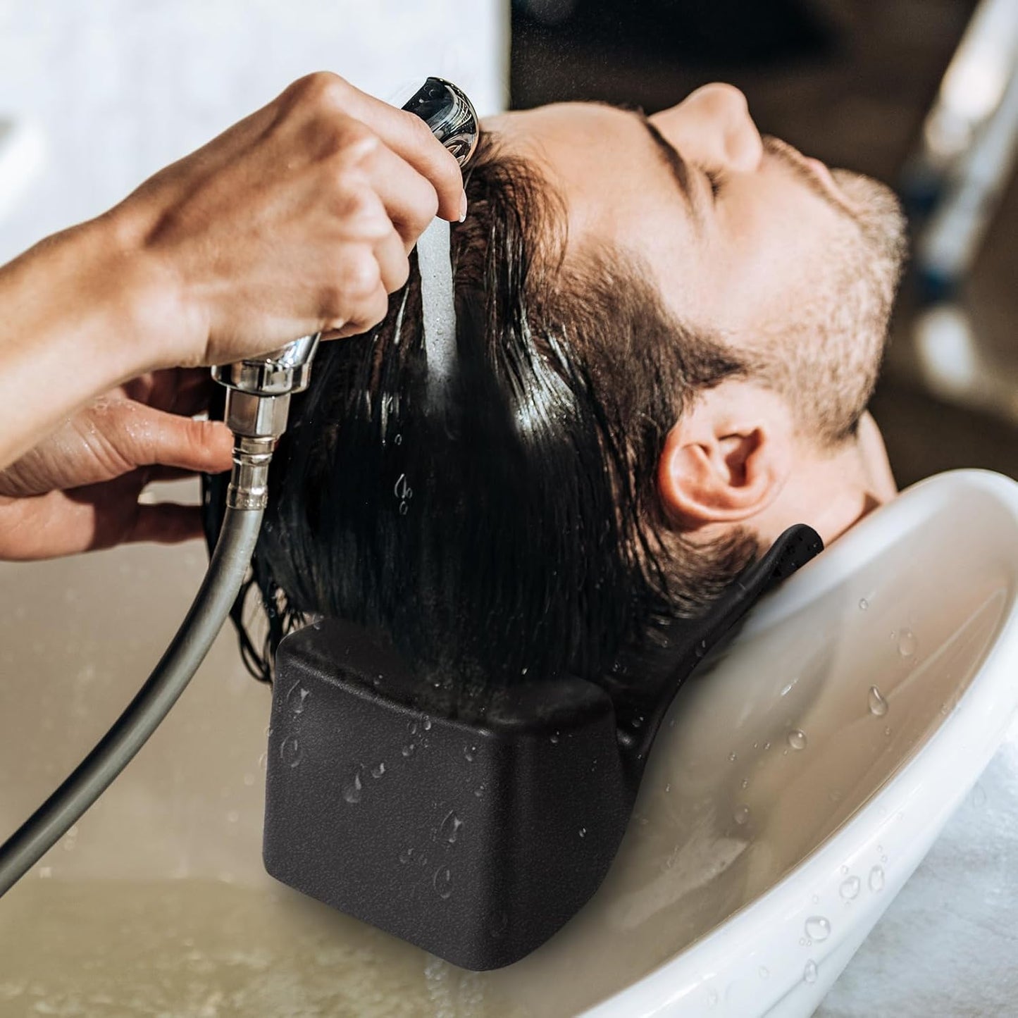 Cadru din salon in care un hairstylist spala parul unui client barbat utilizand suportul negru de protectie pentru gat pe marginea scafei.